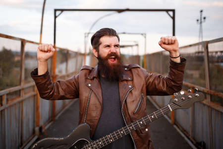 Crazy bearded metalhead with big long mustache in brown leather jacket and jeans holds thumbs up and...