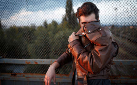 A grown male biker with a long beard, mustache and gray hair in a brown leather jacket leaned his...