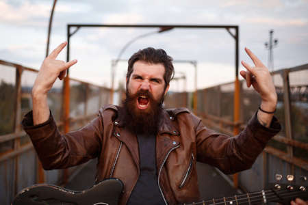 Crazy bearded metalhead with big long mustache in brown leather jacket and jeans holds thumbs up and...