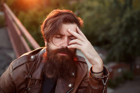 A grown male biker with a long beard, mustache and gray hair in a brown leather jacket leaned his face against an iron mesh fence at sunset.の写真素材
