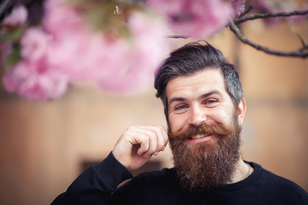 A bearded male programmer with a long beard and dark gray hair in a shirt and jeans squatted on the green grass in the park with a laptop. Game developer resting in natureの写真素材