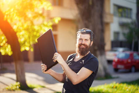 Cheerful bearded male developer with a long beard and dark gray hair in a shirt and jeans hugs a laptop on the background of a green park in springの写真素材