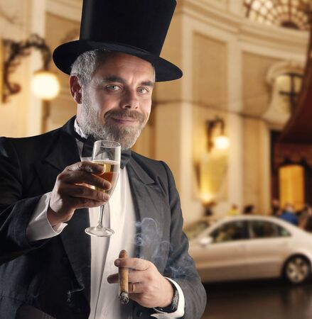 a man wearing a smoking holding a glass and cigar, against a blurry backgroundの写真素材