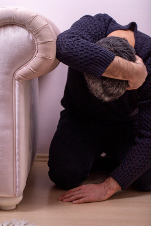 Man protected from earthquake. Frightened man hiding next to sofa in living room during earthquakeの写真素材