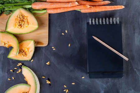 Slices of melon and bunch of carrots on cutting board on black board with notebook and pencil