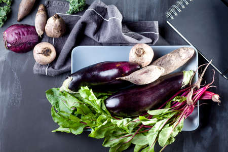 Beetroots, aubergines and beet leaves on the plate with black notebook and cloth on the side, top viewの写真素材