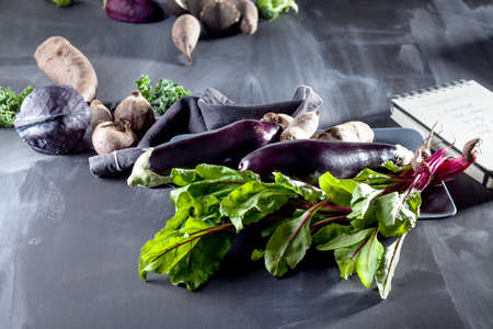 Beetroots, aubergines and beet leaves on the plate with black notebook and cloth on the sideの写真素材