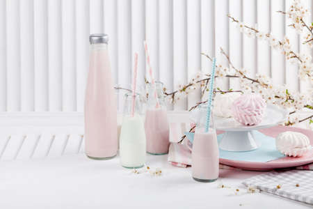 Four bottles of milkshakes, three with straws and plates with pink and white meringues and cloth on white wooden shutter with blossom apple tree in the backgroundの写真素材
