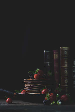 Pancakes in the rusty pan with strawberries and blackberries, honey and old books in backgound with...
