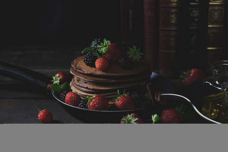 Pancakes in the rusty pan with strawberries and blackberries, honey,fork and old books in backgound with mystical lightの写真素材