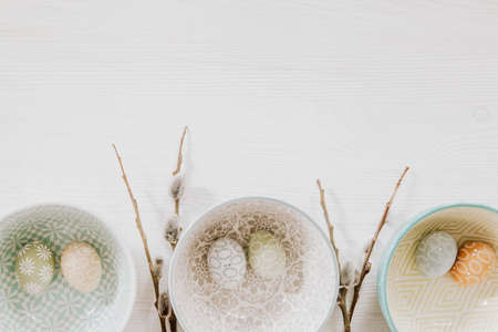 Beautiful pastel eggs in decorative bowls and fluffy willow on the bottom of white wooden backgroundの写真素材