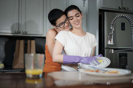 A short hair gay woman embracing her couple while she cleans the dishes. LGBT women in everyday...