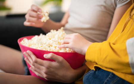 Closeup of the Hands of Two Teenage Girls Eating Pop Corn From a Pink Bowl Sitting on Couch While Watching a Movie Film. Concept of Low Fat Snacks and Good Sources of Dietary Fibers. High sodium foodの写真素材