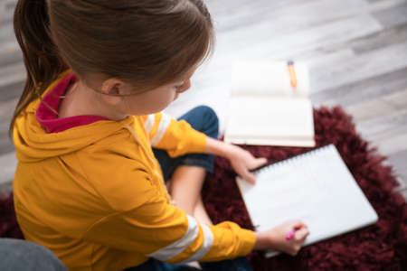 Focused Smart Little Girl Writing with Her Pen and Studying her School Homework while Sitting on The Floor at Home. Concept of Young People, Educationの写真素材