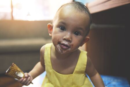 Child eating ice cream / Funny Asian baby girl happy holding ice cream cone waffle with dirty face and mouthの写真素材