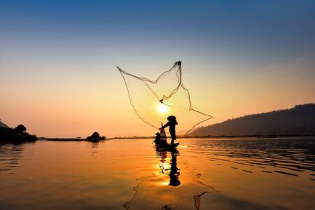 Asia fisherman net using on wooden boat casting net sunset or sunrise in the Mekong river - Silhouette fisherman boat with mountain background life person countrysideの写真素材