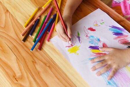Girl painting on paper sheet with colour pencils on the wooden table at home / child kid doing drawing picture and colorful crayonの写真素材
