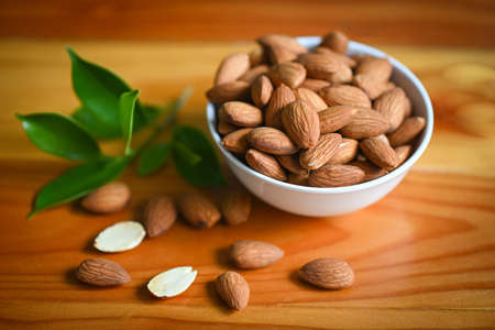 Delicious sweet almonds on the wooden table, roasted almond nut for healthy food and snack, Almonds nuts on white bowl and green leaf on backgroundの写真素材