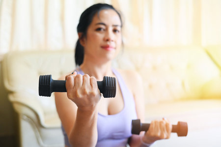 sporty woman exercise muscles in sportswear with dumbbell at home in the living room, woman exercising at home, woman dumbbells in her hands, sport recreation health care conceptの写真素材
