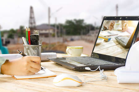 Woman architect working and equipment such as white helmet and house plans on table with background of site construction.の写真素材