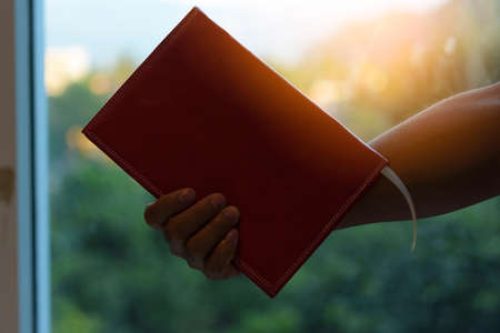Hand holding book in the glass room with sunset background.