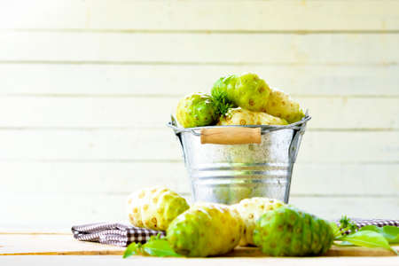 Noni fruit in zinc bucket and noni on table on white wooden background.