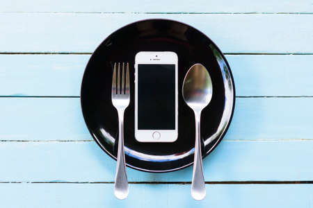 Smartphone on black plate and spoon with fork on blue background.の写真素材