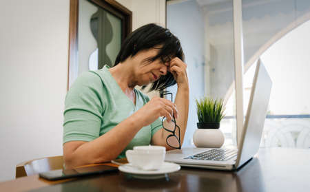 Stressed mature older woman working with computer laptop at home indoor. Smart working senior people and technology.の写真素材