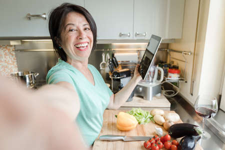 Senior woman taking a selfie cooking vegetables with help of recipe on tablet in the kitchen at homeの写真素材