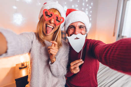 Happy couple taking funny selfie celebrating Christmas time at home - Winter holiday concept with...