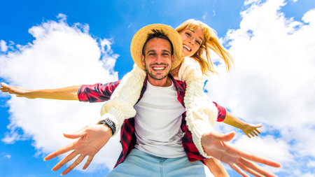 Positive couple having fun outside doing piggyback holding hands fly - Happy boyfriend and girlfriend smiling at camera standing on a blue sky backgroundの写真素材