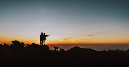 Happy couple with dog embracing while standing on the top of the hill looking at sunsetの写真素材