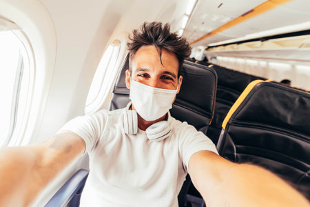 Young man wearing protective face mask taking selfie portrait sitting on the aircraft seat near the windowの写真素材