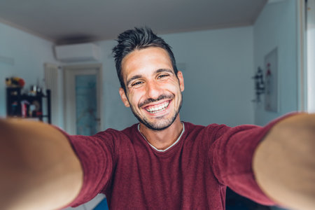 Happy young handsome millennial guy taking a selfie smiling at the camera in the living room at homeの写真素材