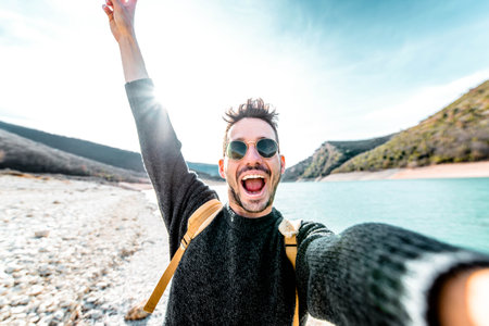 Handsome man with backpack taking selfie in the nature - Male tourist smiling at camera while walking on mountainsの写真素材