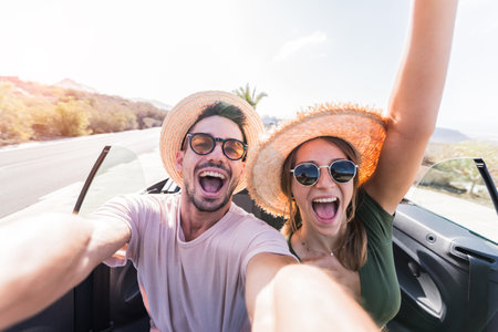 Happy beautiful couple in love taking a selfie portrait driving a convertible car on the road at vacationの写真素材