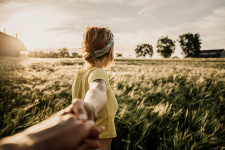 Portrait of a happy young woman holding hand of her boyfriend while walking by a wheat field at sunsetの写真素材