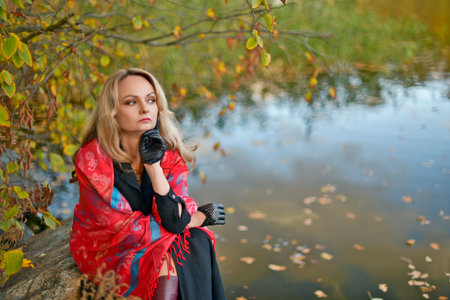 A woman in her prime, dressed in a formal black dress, a red headscarf, and a large red umbrella, sits along the riverbank in a colorful autumn forestの写真素材