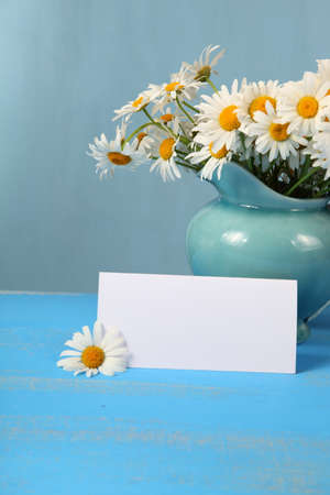 Beautiful chamomiles in a blue jug and greeting card on wooden tableの写真素材