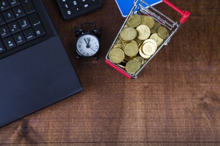Shopping trolley with coins,  laptop and alarm clock on a table.Workplace of  businessman.の写真素材