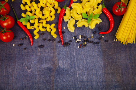 Different pasta on a dark wooden background. Raw pasta, chili, tomatoes and basil close-up.の写真素材