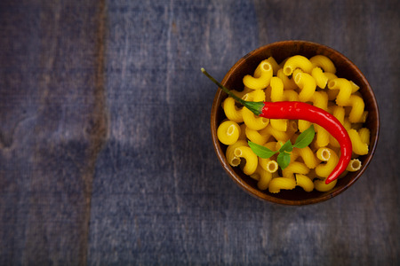 Pasta in a wooden bowl with hot chili and basil on a dark wooden background. Raw pasta close-up.の写真素材