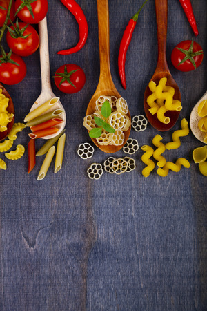 Different pasta in spoons on a dark wooden background, top view. Raw pasta, chili, tomatoes and basil close-up.の写真素材