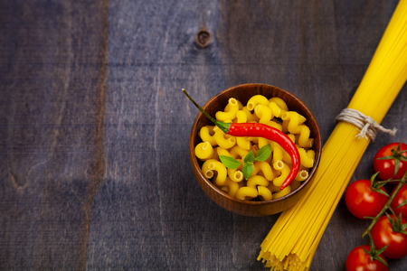 Different pasta on a dark wooden background. Pasta in a wooden bowl, spaghetti, tomatoes and basil close-up.の写真素材