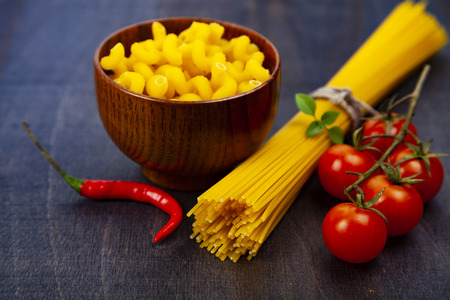 Different pasta on a dark wooden background. Pasta in a wooden bowl, spaghetti, tomatoes and basil close-up.の写真素材