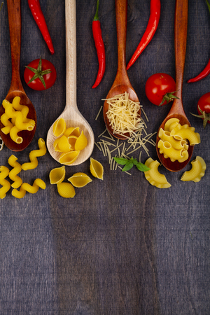 Different pasta in spoons on a dark wooden background, top view. Raw pasta, chili, tomatoes and basil close-up.の写真素材