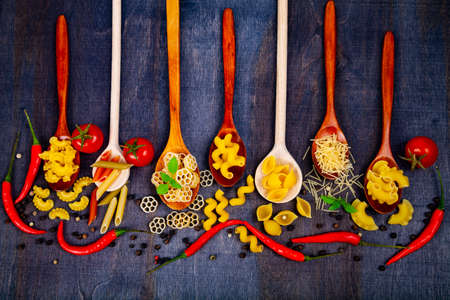 Different pasta in spoons on a dark wooden background, top view. Raw pasta, chili, tomatoes and basil close-up.の写真素材