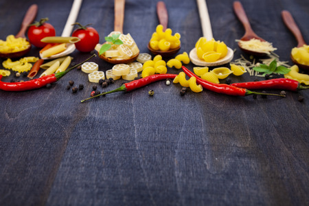 Different pasta in spoons on a dark wooden background. Raw pasta, chili, tomatoes and basil close-up.の写真素材