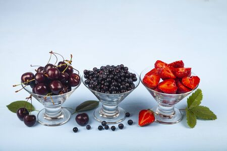 Ripe berries in transparent bowls on a blue wooden background. Blueberries, cherries and...