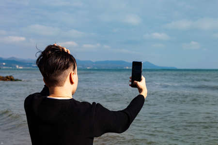 Young guy in a black jacket with a phone sits on the seashore. Man takes a selfie near the sea.の写真素材